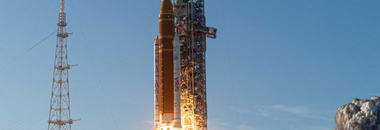 Rocket on a launch pad beside a tall support tower, with flame and smoke at the base against a clear blue sky.